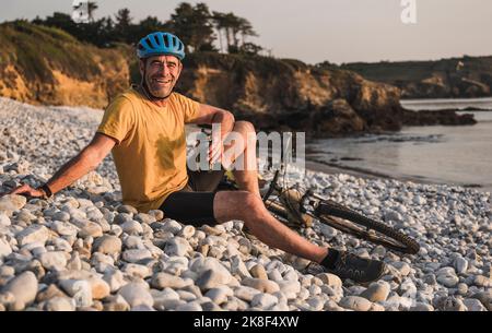 Glücklicher Mann, der am Fahrrad mit einer Wasserflasche am Strand sitzt Stockfoto