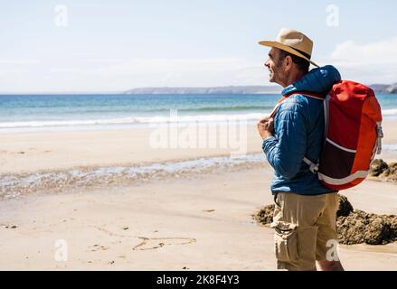 Mann mit Rucksack, der Hut trägt und am sonnigen Tag am Strand steht Stockfoto