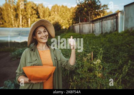 Lächelnde, reife Frau mit einer Schale aus grünen Erbsen, die die Daumen nach oben im Garten gestikuliert Stockfoto