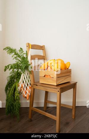 Netzbeutel mit Karotten und Kiste mit Kürbissen auf dem Stuhl vor der Wand Stockfoto