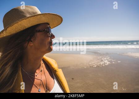 Reife Frau mit Hut und Sonnenbrille am Strand an sonnigen Tagen Stockfoto