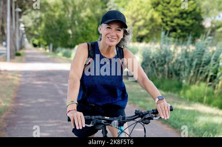 Glückliche ältere Frau trägt Mütze Fahrrad auf dem Fußweg fahren Stockfoto