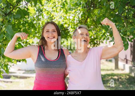 Fröhlicher Fitnesstrainer, der Muskeln anbiegt und eine ältere Frau vor dem Baum steht Stockfoto