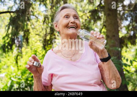 Glückliche ältere Frau mit Wasserflasche, die vor dem Baum im Park steht Stockfoto
