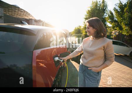 Glückliche Frau, die an einem sonnigen Tag am Straßenrand ein Elektroauto lädt Stockfoto