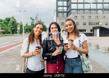 Lächelnde multirassische Freunde mit wiederverwendbaren Kaffeetassen, die auf dem Fußweg stehen Stockfoto