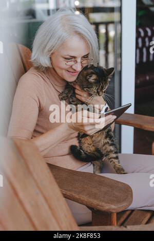 Glückliche, reife Frau, die mit einer Katze sitzt und einen Tablet-PC zu Hause benutzt Stockfoto
