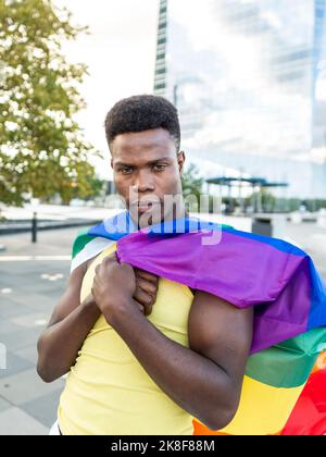 Selbstbewusster junger Mann, eingewickelt in Regenbogenfahne auf dem Fußweg Stockfoto