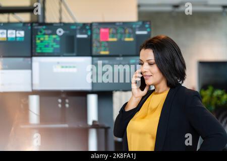 Lächelnde Geschäftsfrau, die auf dem Mobiltelefon vor Computermonitoren spricht Stockfoto