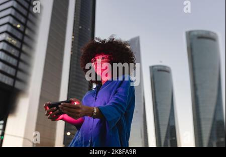 Rotes Licht fällt auf das Gesicht einer Frau, die eine Smartbrille trägt und das Smartphone vor dem Gebäude hält Stockfoto