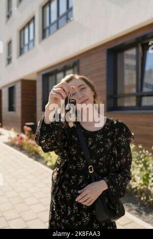 Lächelnde junge Frau, die Hausschlüssel vor dem Gebäude zeigt Stockfoto