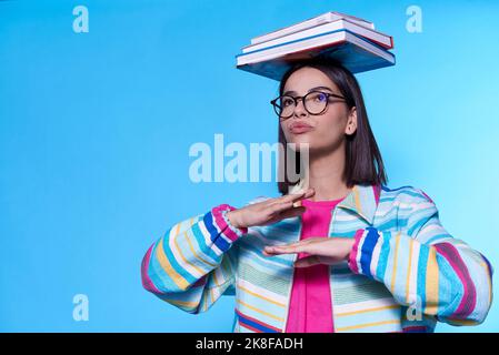 Junge Frau trägt bunte Reißverschluss balancierende Bücher auf dem Kopf vor blauem Hintergrund Stockfoto