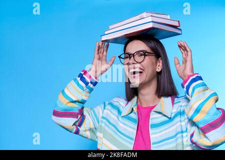 Glückliche junge Frau trägt bunte Reißverschluss balancierende Bücher auf dem Kopf vor blauem Hintergrund Stockfoto