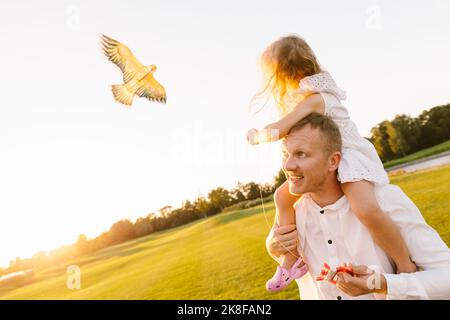 Vater trägt Tochter auf Schulter fliegenden Drachen im Park Stockfoto