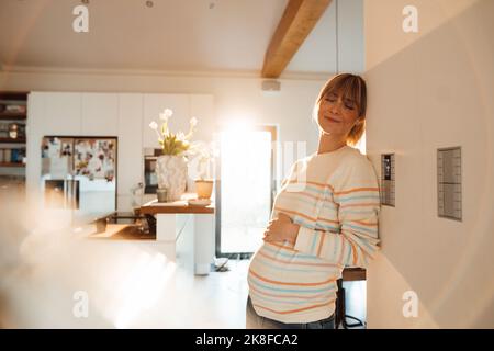 Erwartungsvolle Frau mit geschlossenen Augen, die sich zu Hause am Thermostat an der Wand lehnt Stockfoto