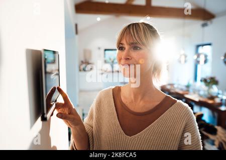 Lächelnde Frau, die die Smart-Home-App über einen Tablet-PC nutzt, der zu Hause an der Wand montiert ist Stockfoto