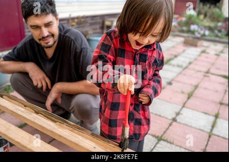 Lächelnder Mann, der einen glücklichen Sohn ansah, der eine Holzbank malte Stockfoto