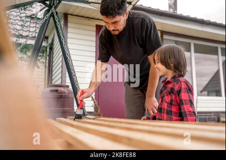 Glücklicher Junge, der den Vater ansieht, der vor dem Haus eine Schaukel auf einer Holzbank malt Stockfoto