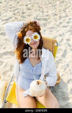 Lächelnde Frau mit Blumensonnenbrille sitzt mit Kokosnuss am Strand Stockfoto