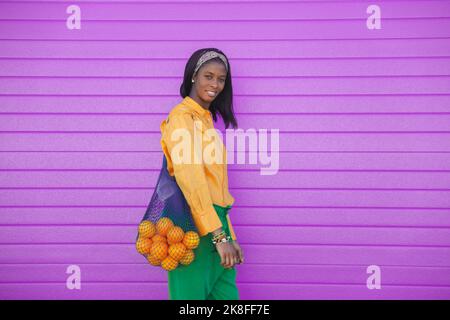 Lächelnde junge Frau, die mit einem Beutel mit Orangen vor der lila Wand steht Stockfoto