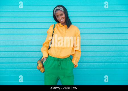 Lächelnde Frau mit einem Beutel mit Orangen, die vor der blauen Wand stehen Stockfoto