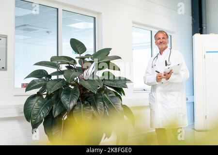 Lächelnder Oberarzt mit Zwischenablage, der neben einer Topfpflanze steht Stockfoto