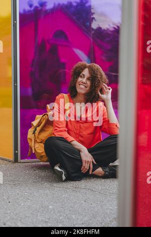 Lächelnde Frau mit Hand im Haar, die mit gekreuzten Beinen durch eine bunte Brille sitzt Stockfoto