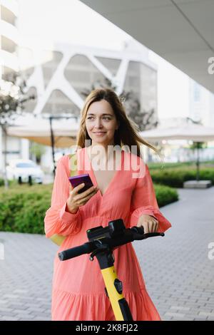 Lächelnde Frau hält Smartphone stehen mit Elektro-Roller auf dem Fußweg Stockfoto