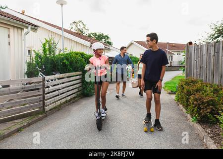 Bruder mit Skateboard Blick auf Schwester Reiten Elektroroller auf der Straße Stockfoto