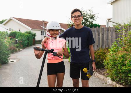 Lächelnder Junge mit Arm um die Schwester, der Helm trägt und auf der Straße steht Stockfoto