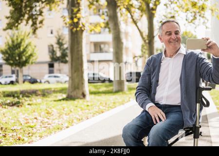 Lächelnder älterer Mann mit Videoanruf über das Smartphone, der auf einem Gehwagen sitzt Stockfoto