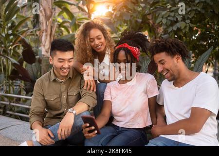 Junge Frau, die ein Smartphone mit glücklichen Freunden teilt, die an der Wand sitzen Stockfoto