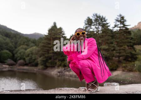 Glückliche junge Frau mit Sonnenbrille, die vor dem See hockt Stockfoto