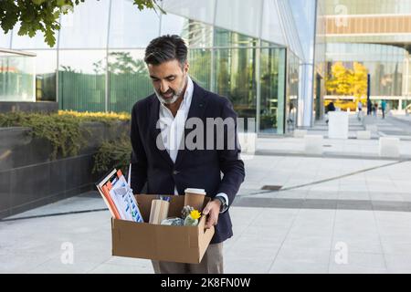 Trauriger Geschäftsmann, der Bürobedarf in einem Kasten neben dem Gebäude ansieht Stockfoto