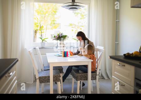 Mutter und Sohn verwenden Farbtuben in Malbuch auf Esstisch zu Hause Stockfoto