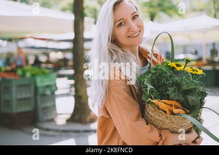 Lächelnde Frau mit Blumen und Gemüse in wiederverwendbarem Weidensack auf dem Markt Stockfoto