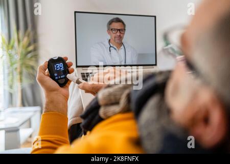 Ein Mann, der den Blutzuckerspiegel über das Gerät überprüft, führt einen Videoanruf mit dem Arzt auf dem Laptop durch Stockfoto