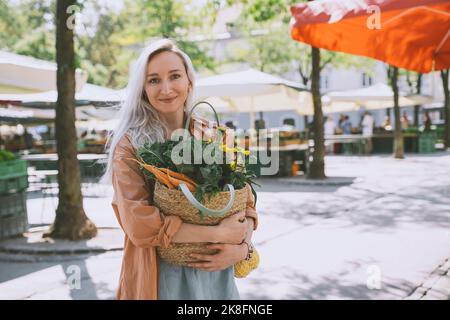 Lächelnde Frau, die einen Beutel mit Gemüse und Blumen auf dem Markt hält Stockfoto