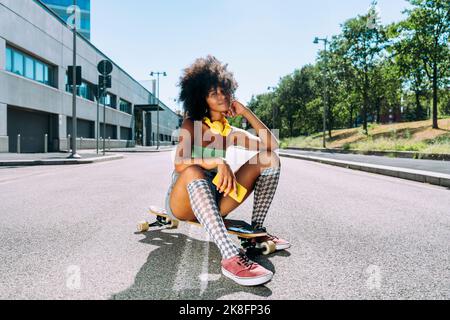 Lächelnde Frau, die auf dem Skateboard auf der Straße sitzt Stockfoto