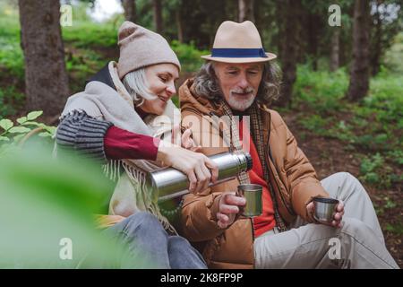 Reife Frau, die Wasser aus Thermoskannen von einem älteren Mann im Wald gießt Stockfoto