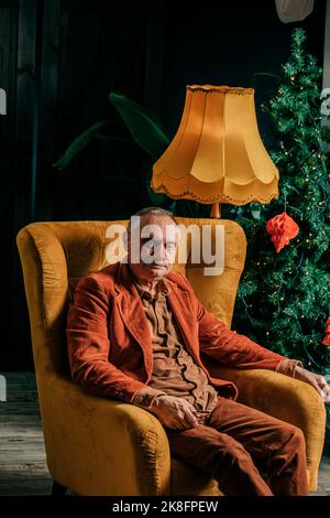 Nachdenklicher älterer Mann, der zu Hause im Sessel am Weihnachtsbaum sitzt Stockfoto