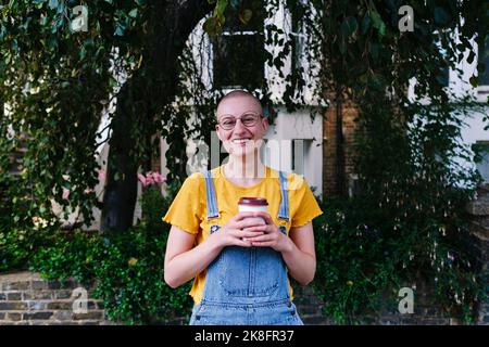 Glückliche nicht-binäre Person mit Einweg-Kaffeetasse vor Pflanzen Stockfoto