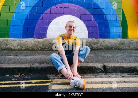 Fröhliche Transgender-Person, die auf einem Fußweg vor der Regenbogenwand sitzt Stockfoto
