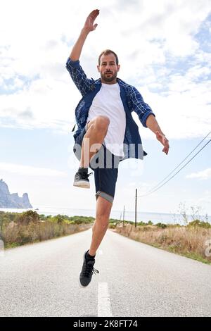 Mann mit erhobener Hand springt auf der Straße vor bewölktem Himmel Stockfoto