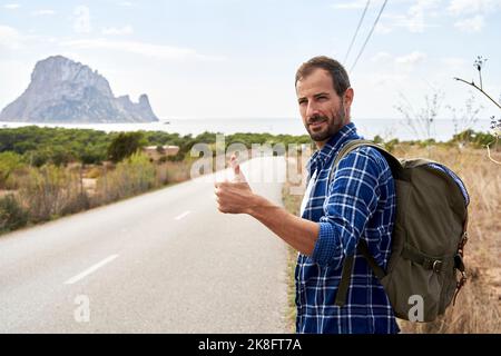 Lächelnder Mann im Rucksack, der den Daumen nach oben auf der Straße zeigte Stockfoto