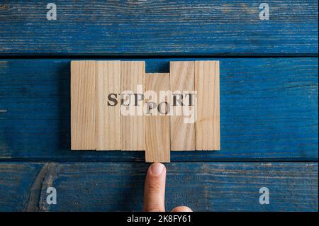 Männlicher Finger, der ein Schild mit der Aufschrift „Support“ auf hölzernen Pflöcken montiert, das auf rustikalen blauen Holzbrettern angebracht ist. Stockfoto