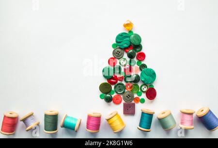 Ein Weihnachts- oder Apfelbaum aus Knöpfen in grüner, roter Farbe. Mehrfarbige Fäden mit Holzspulen. Weißer Hintergrund, Kopierbereich. Kreatives Konzept. Stockfoto