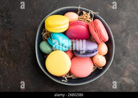 Bunte Makronen und Schokoladeneier in Keramikschale auf schwarzem Beton-Hintergrund. Draufsicht, flach liegend, Nahaufnahme, Stillleben. Frühstück, Morgen, Stockfoto