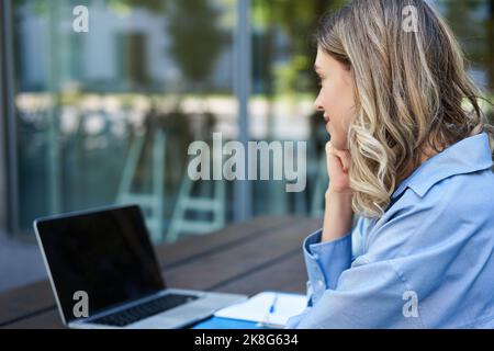 Nahaufnahme eines Porträts einer Studentin, die an Online-Kursen teilnimmt, mit einem Laptop an der frischen Luft sitzt und sich Notizen macht. Videochat für Geschäftsfrau Stockfoto