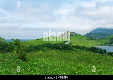 Natürliche Landschaft der Insel Kunashir mit grasbewachsenen Hügeln, vulkanischen Felsen, Vulkan in den Wolken und einem Tal mit einer Lagune Stockfoto